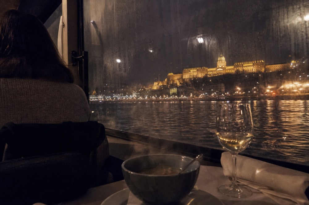Dinner cruise table setting with view of Buda Castle and the Danube at night, no text, no logos, no people