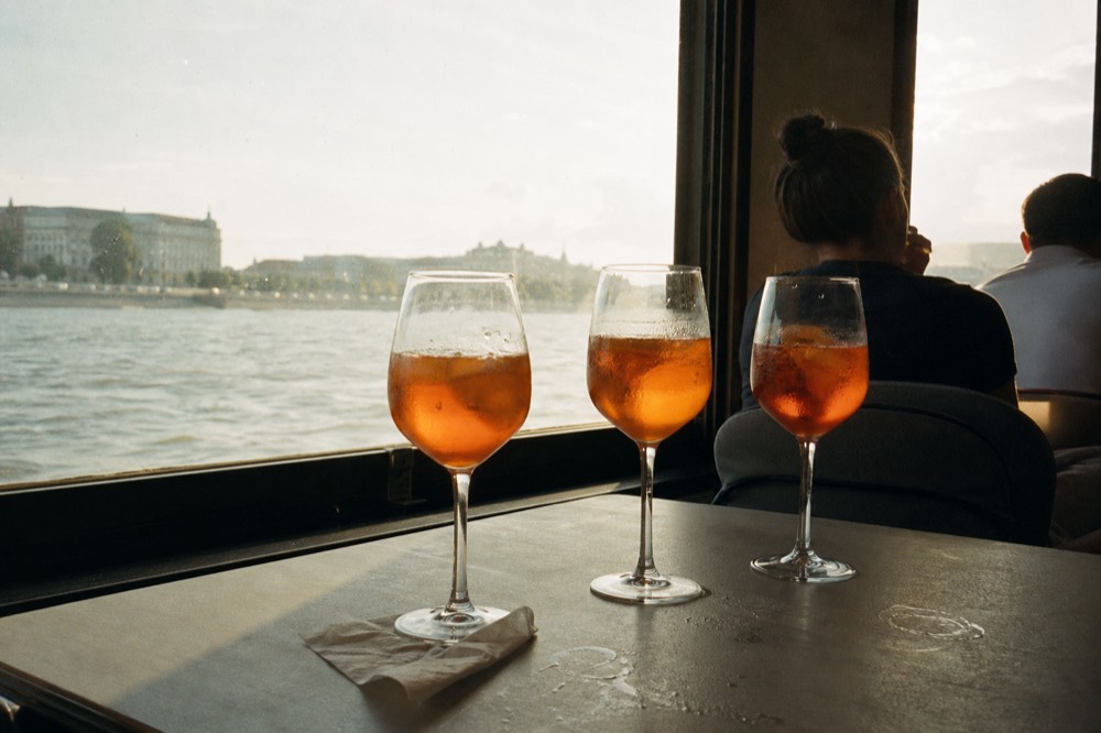 Glasses of orange spritz drinks on a boat table with river views behind, no text, no logos, no people