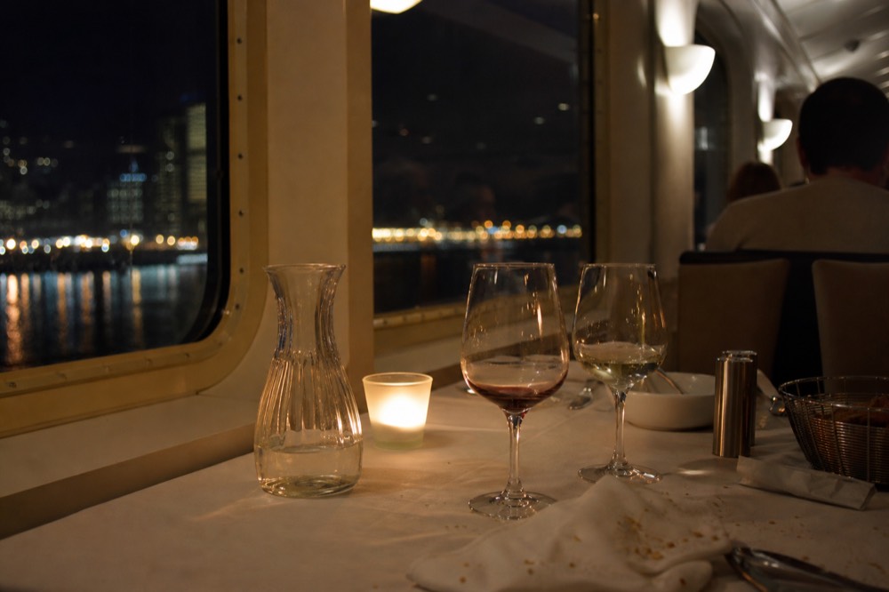 Passengers at a candlelit dinner table aboard a Danube cruise boat with the city lights outside the window, no text, no logos, no people