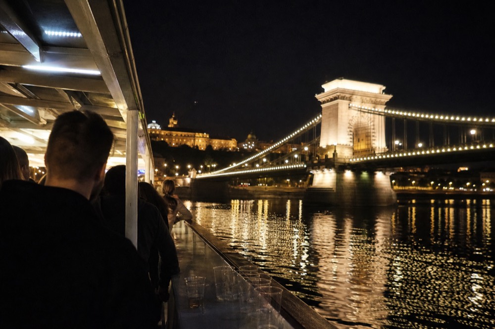 A drinks cruise boat with passengers on the open deck passing the illuminated Chain Bridge at night, no text, no logos, no people