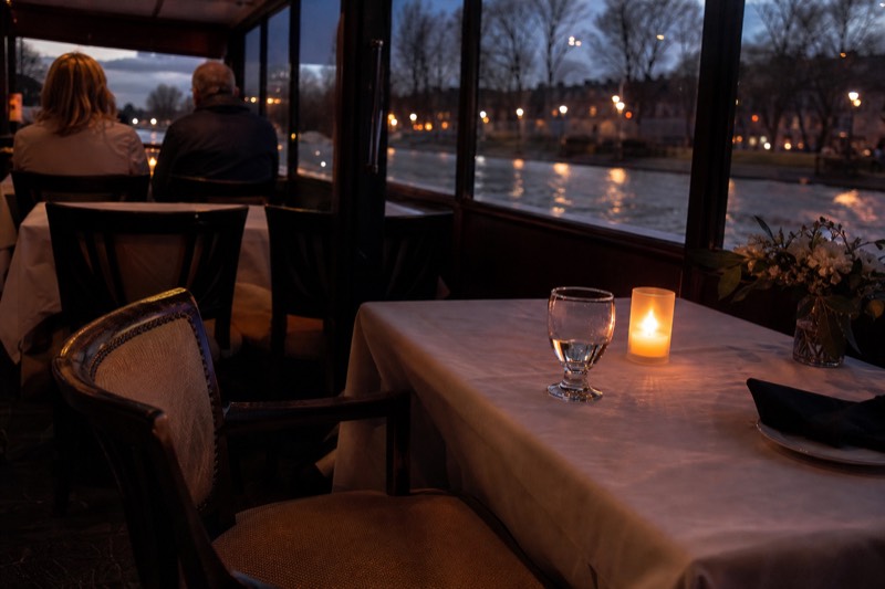 A table set on a glass-enclosed Danube dinner cruise boat with the lit Budapest riverbanks outside