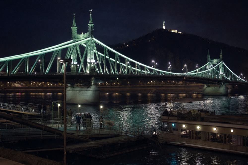 Green Liberty Bridge illuminated over dark Danube water with Gellert Hill behind, no text, no logos, no people