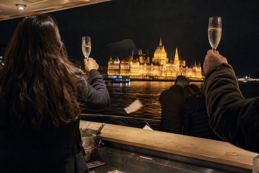 Passengers toasting with prosecco on a Danube night cruise with illuminated Parliament in the background, no text, no logos, no people