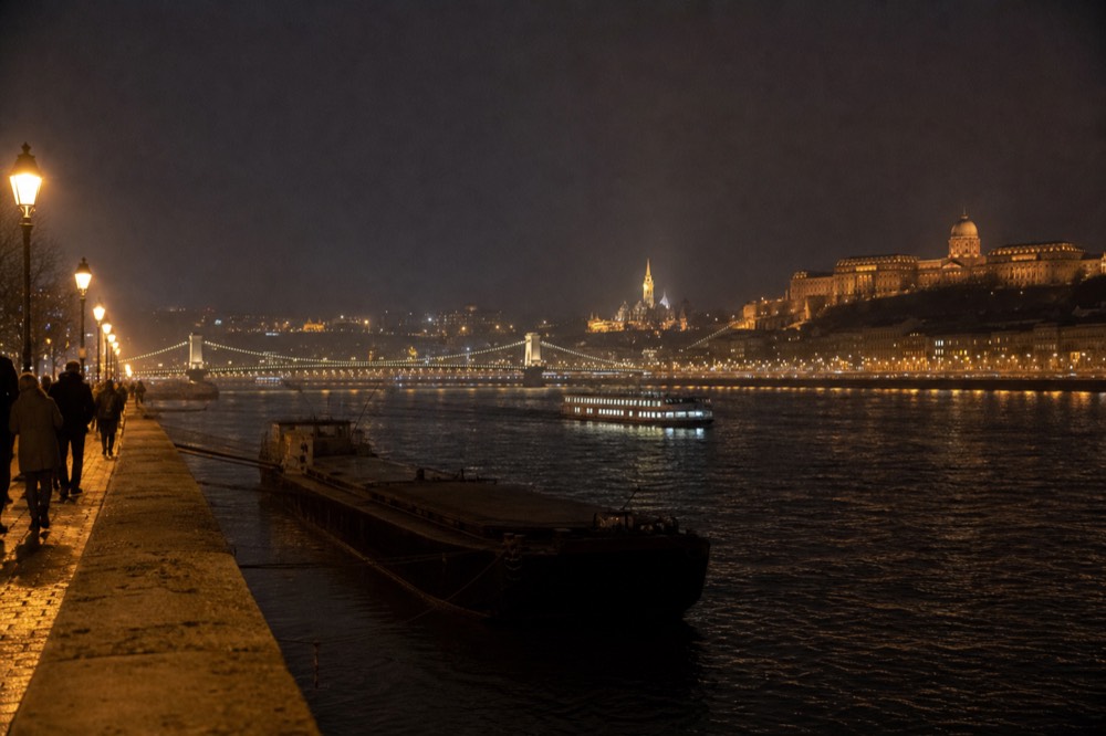 Wide shot of the Danube at night with multiple illuminated landmarks and a cruise boat, no text, no logos, no people