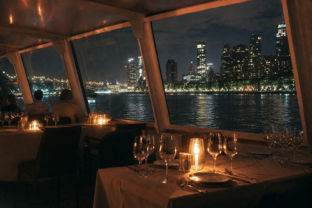 Candlelit tables set for dinner on a Danube cruise boat with the city glowing through panoramic windows, no text, no logos, no people.