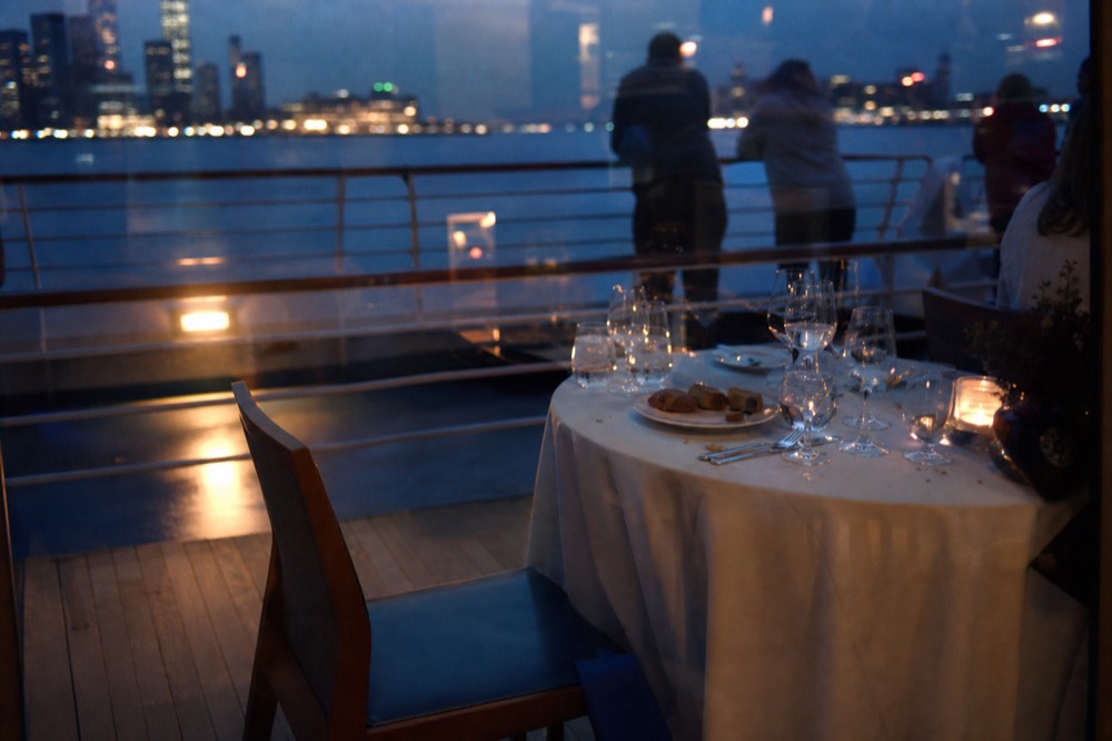 Elegantly set dinner table on a glass-enclosed river cruise boat with city lights outside, no text, no logos, no people