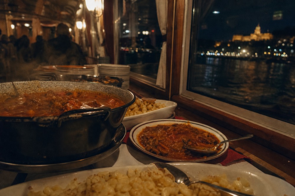 Buffet table on a Budapest dinner cruise boat with Hungarian dishes and city view through windows, no text, no logos, no people
